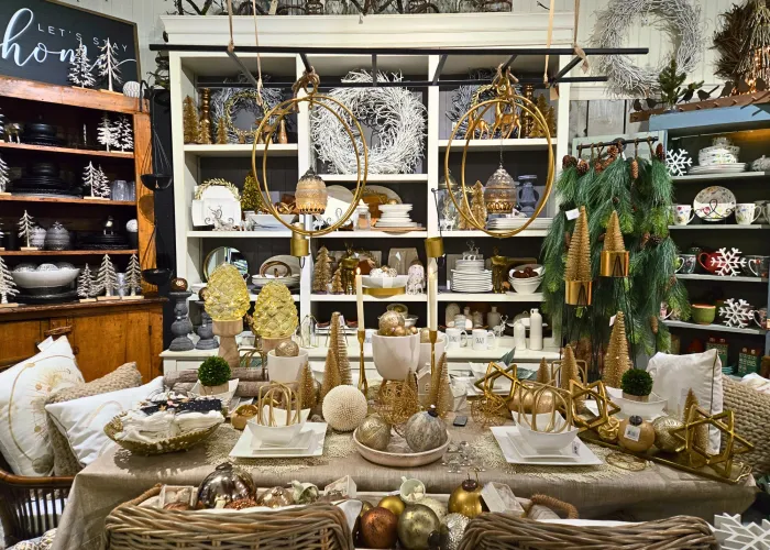 A picture of holiday decorations on a table with greenery and gold accents. Shelves in the background have glassware and decor on display. 